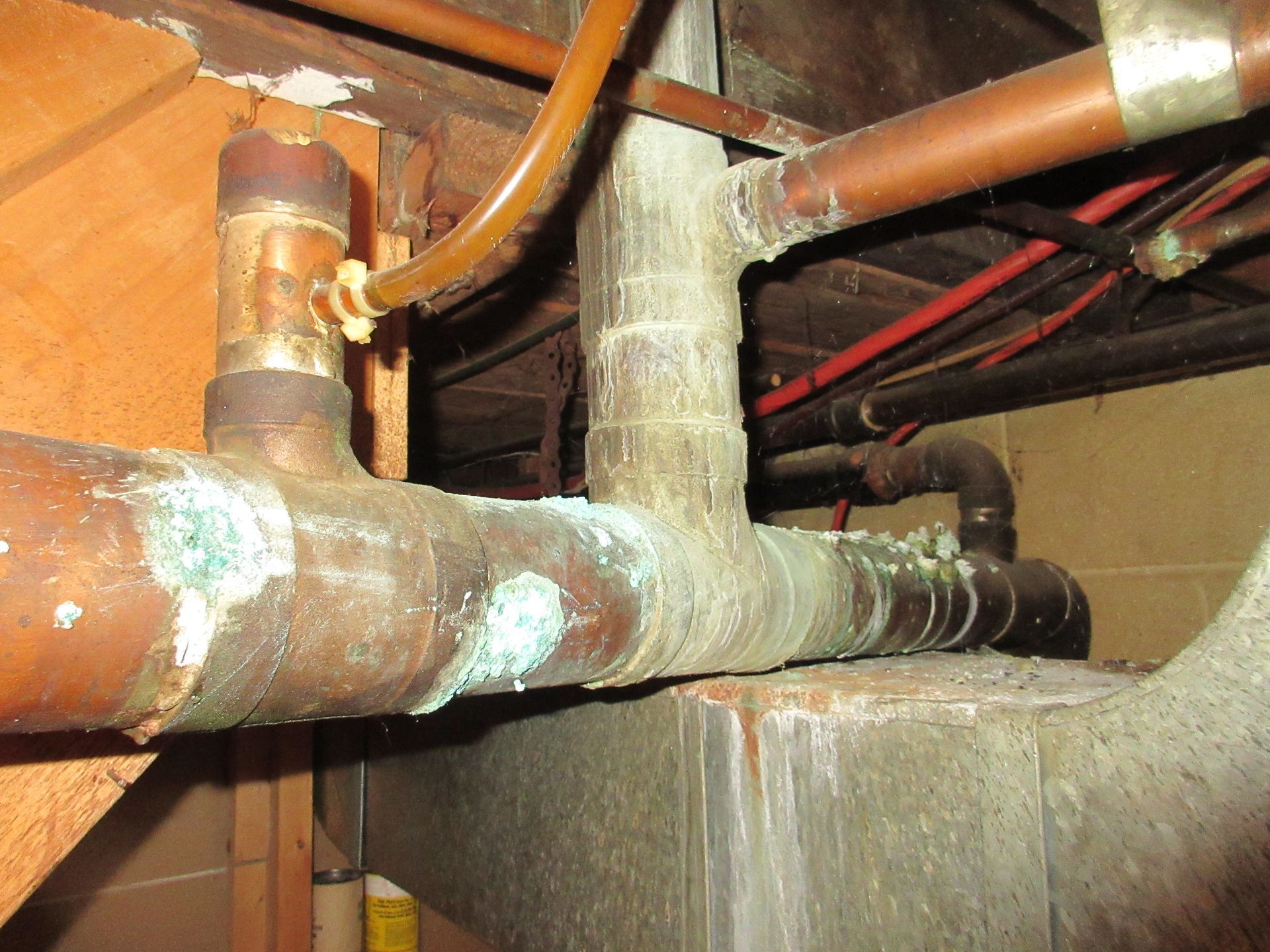 A series of copper pipes under a building, showing significant blue-green corrosion at the joints and connections.