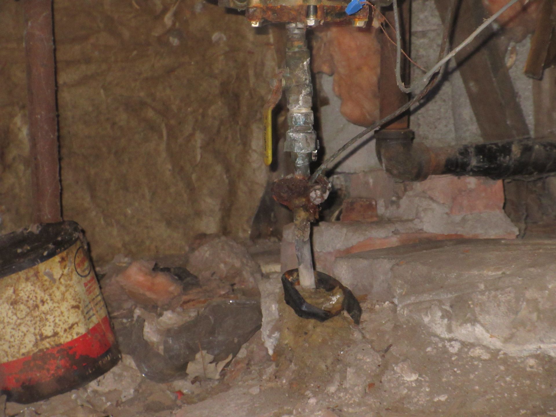 A vertical metal pipe with a valve, partially wrapped in black tape, extends from a dark basement floor near debris.