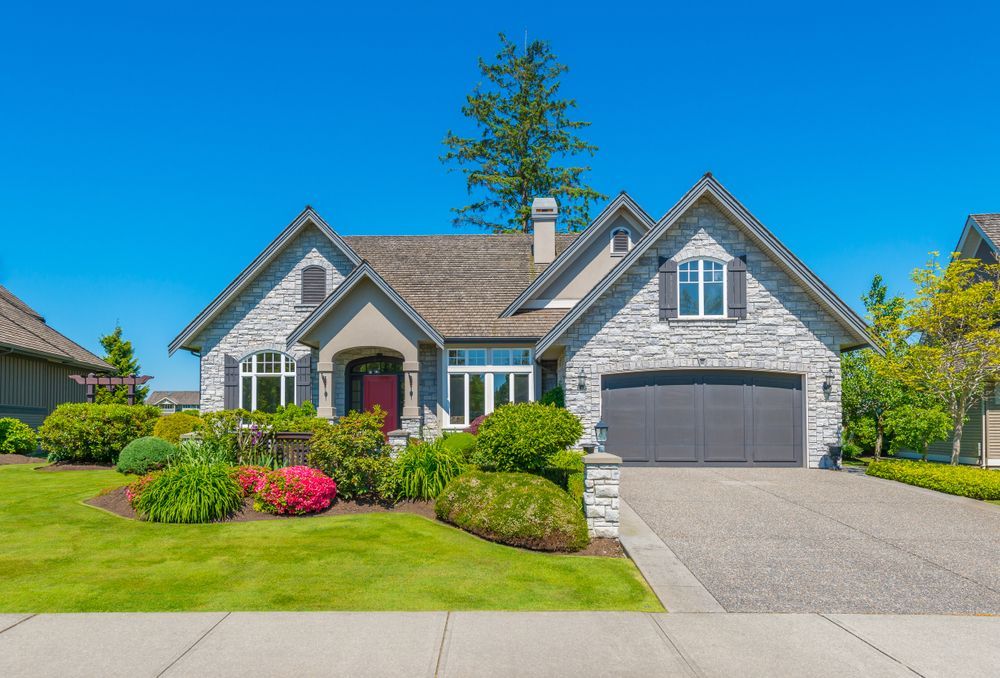 A gray stone single-family home with a two-car garage, manicured lawn, and colorful landscaping under a clear blue sky.