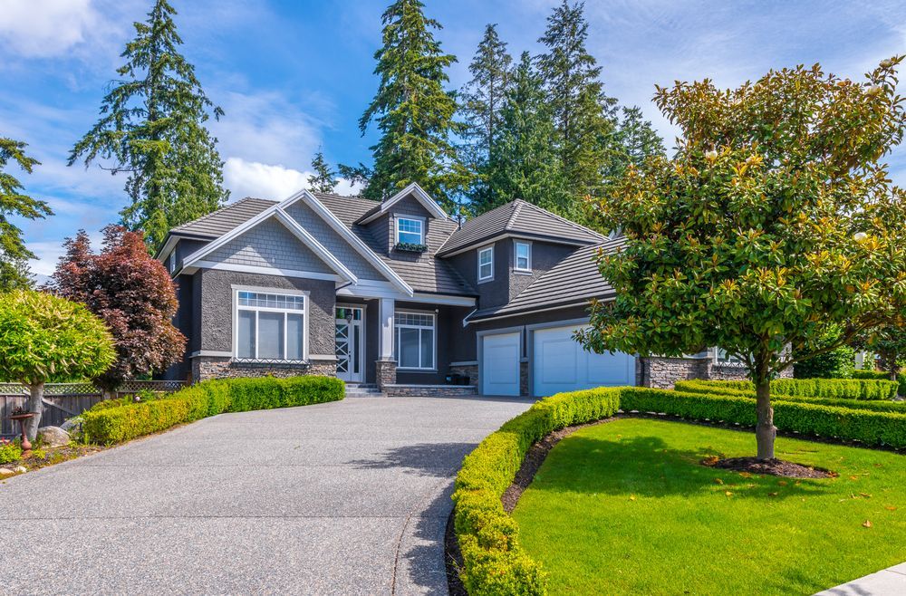 A two-story grey suburban house with a paved driveway, neatly manicured hedges, and a green lawn under a blue sky.
