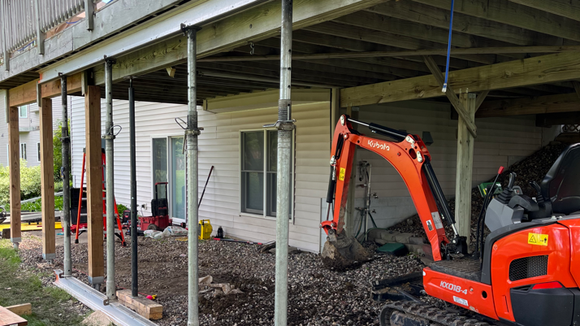 Orange excavator working under a raised house on posts, clearing gravel in a crawl space