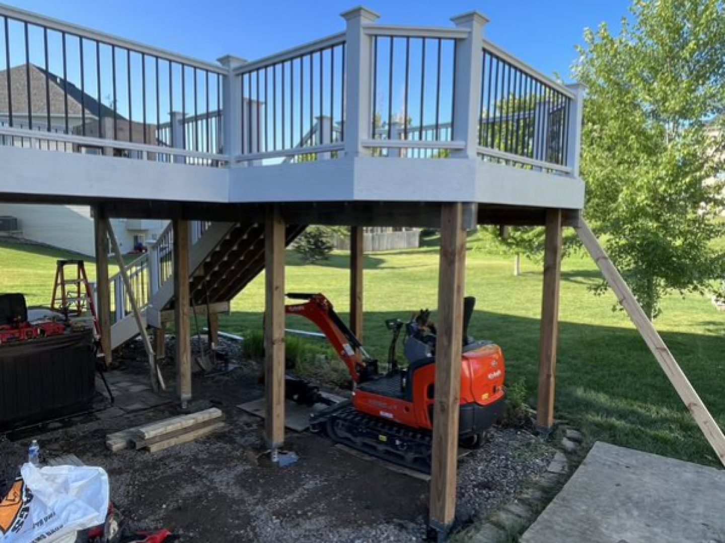 Raised wooden deck under construction with stairs, railings, support posts, and an orange mini excavator below