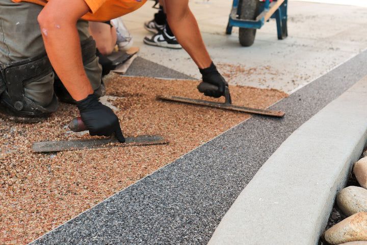 A worker in gloves uses trowels to smooth a mixture of decorative colored pebbles on a concrete patio surface.