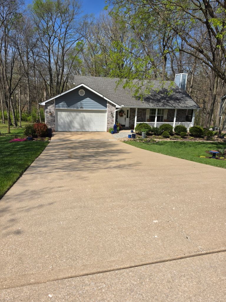 Suburban house with a long driveway, white garage, and front porch surrounded by trees and lawn.