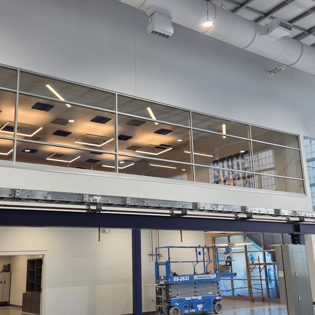 Indoor industrial space with mezzanine windows, ceiling lights, scaffolding, and a blue lift near a white brick wall