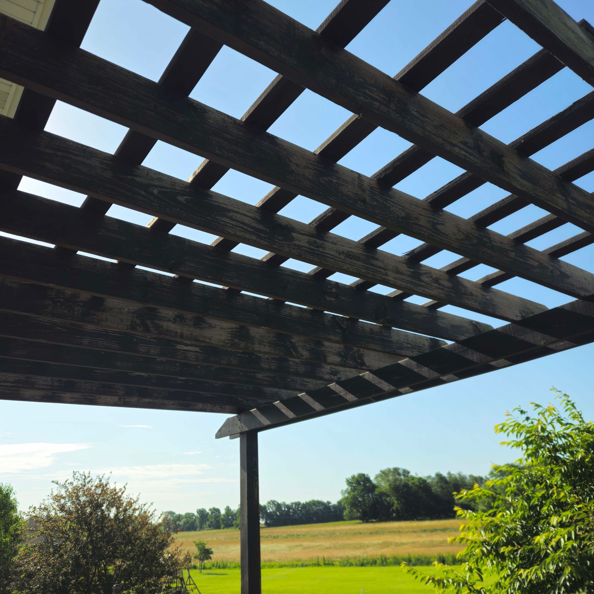 Pergola roof framing with blue sky over a grassy field and trees
