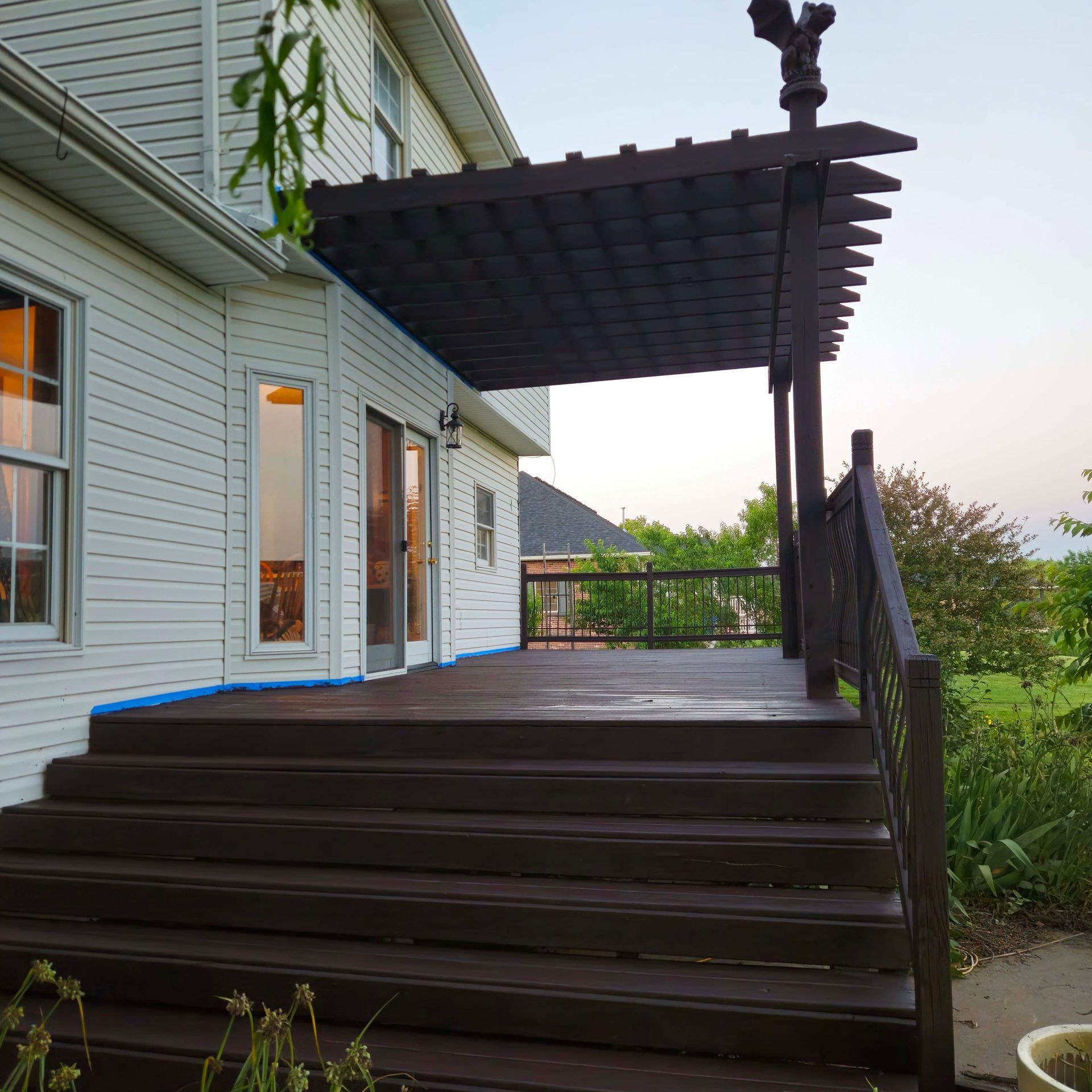 Wooden deck with pergola beside a light gray house, viewed from steps overlooking greenery.