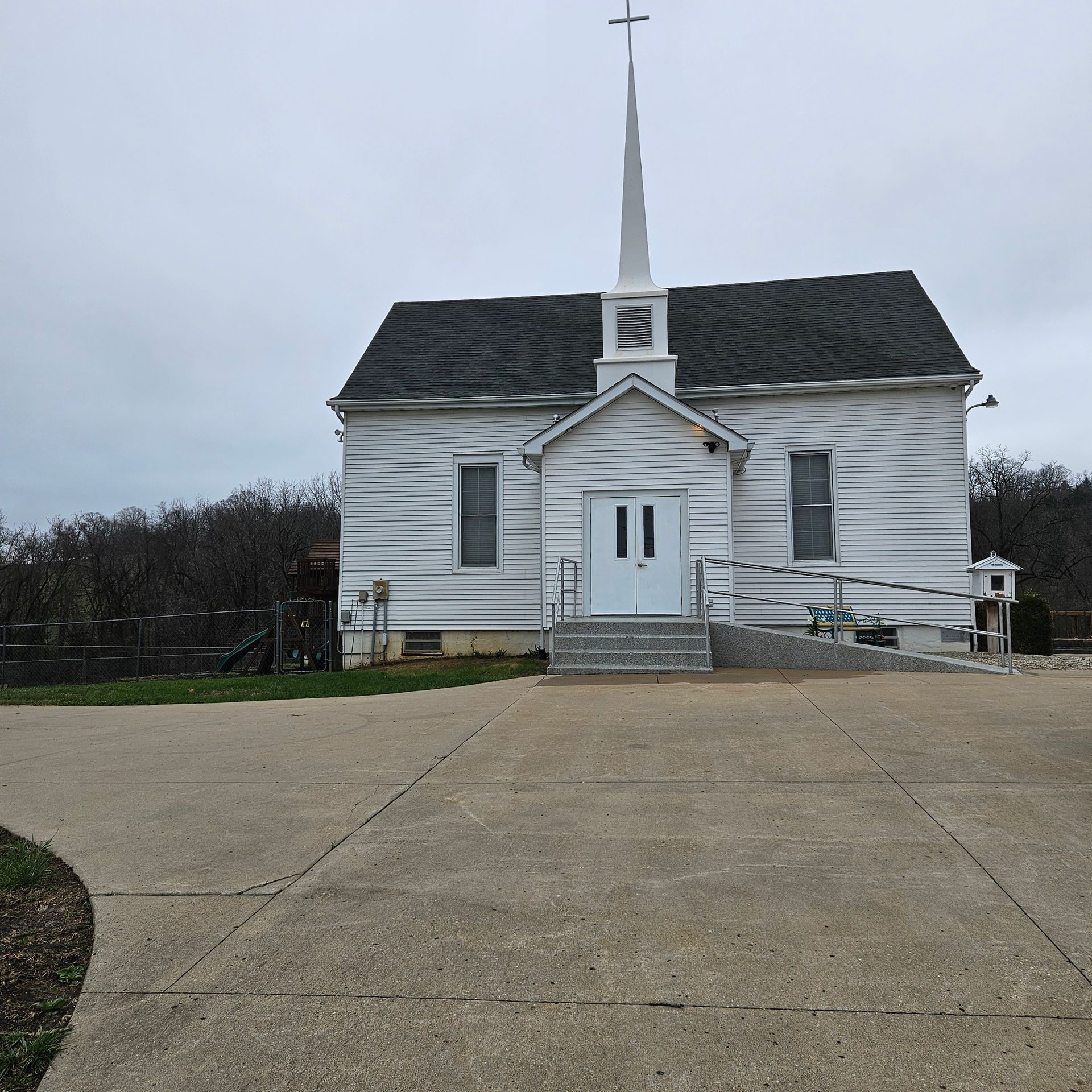 Small white church with steeple and ramp, viewed from a concrete driveway under a cloudy sky.