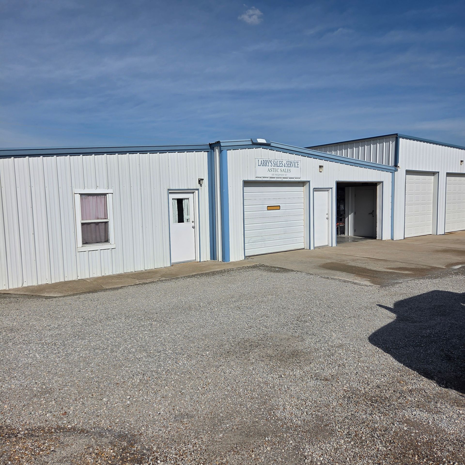 White industrial building with blue trim, garage door, and gravel lot under a clear sky