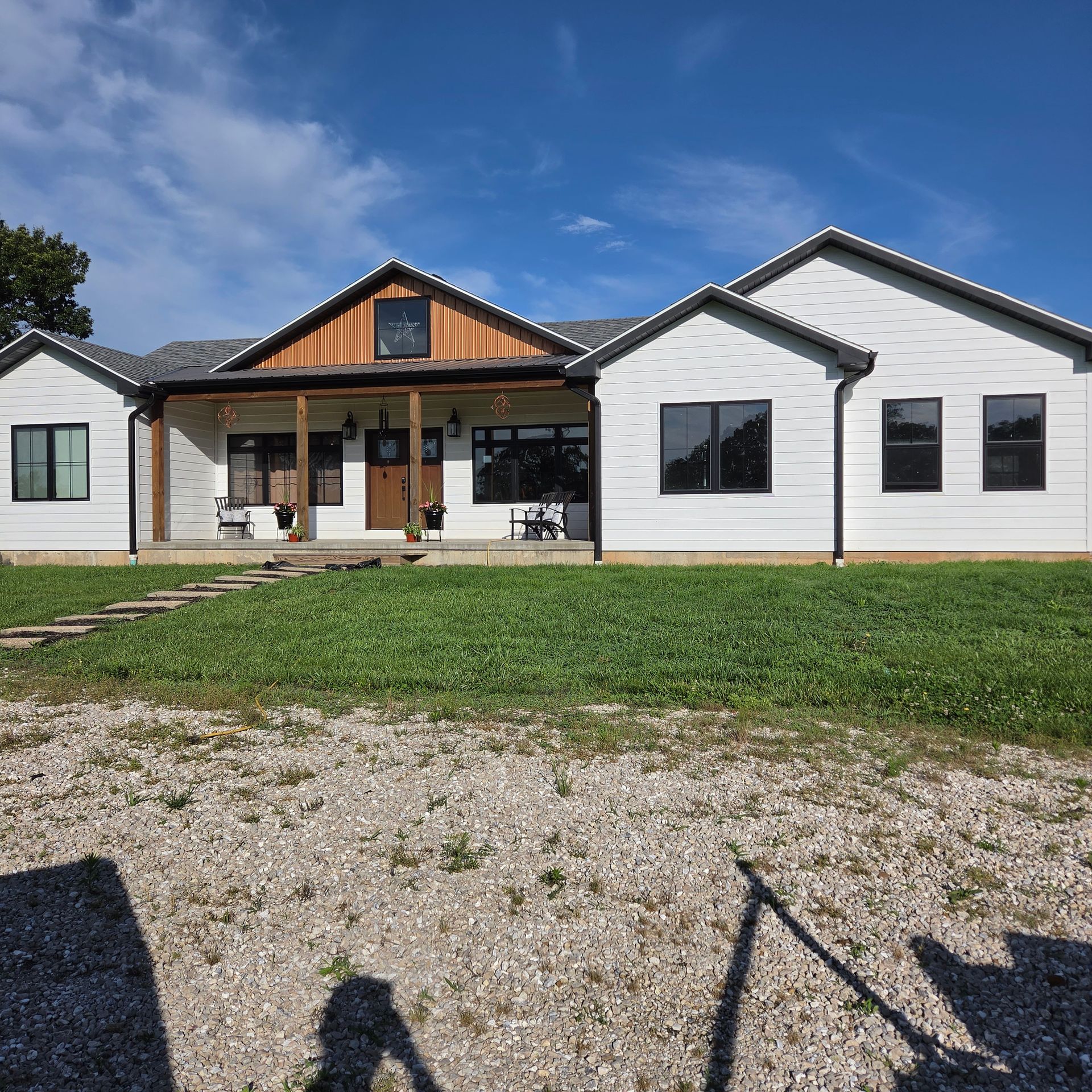 Single-story white house with wood porch accents, black windows, and a gravel driveway under a blue sky
