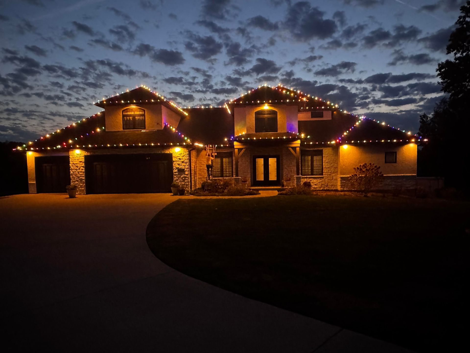 Large house at dusk with warm holiday lights glowing along the roofline and garage.