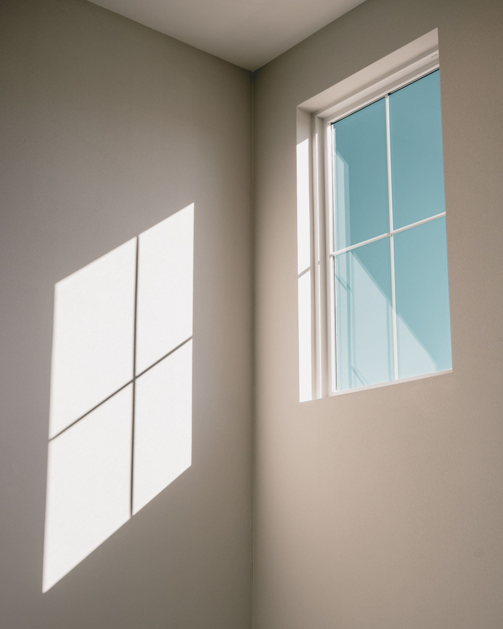 Sunlight casts a window shadow on a beige wall beside a small blue-tinted window.