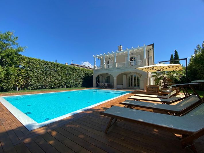Villa soleggiata con piscina turchese, terrazza in legno e lettini prendisole sotto un cielo azzurro limpido.
