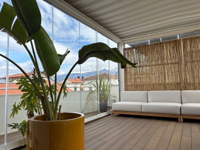 Patio moderno con divano bianco, grande pianta in vaso, pavimentazione in legno e vista sulla città oltre la ringhiera in vetro.