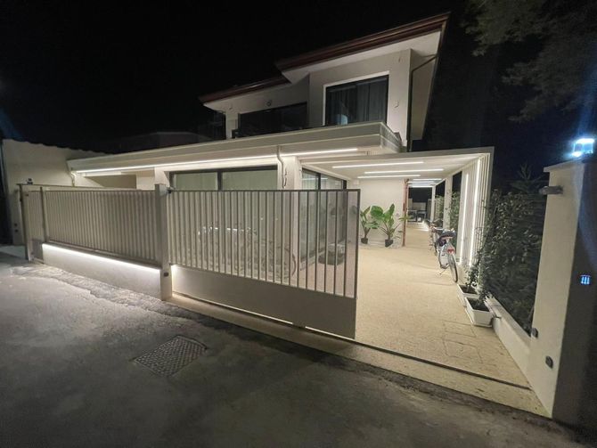 Casa moderna di notte con vialetto illuminato, muri bianchi e cancello in metallo lungo la strada.