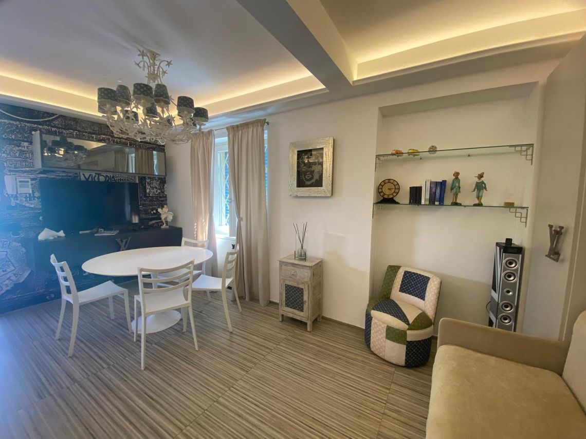 Luminosa sala da pranzo con tavolo rotondo bianco, sedie, divano, mensole e calda illuminazione a soffitto.