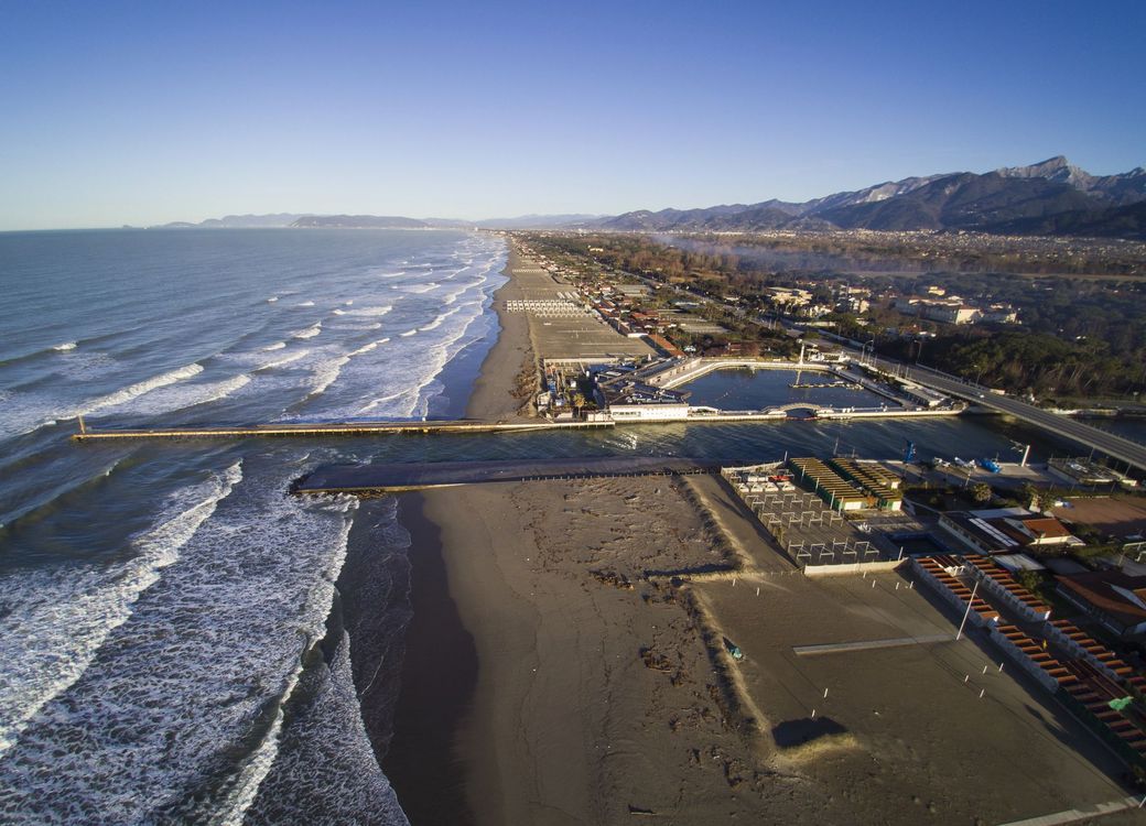 Veduta aerea di una spiaggia costiera di Forte dei marmi con onde, molo ed edifici sul lungomare al tramonto.
