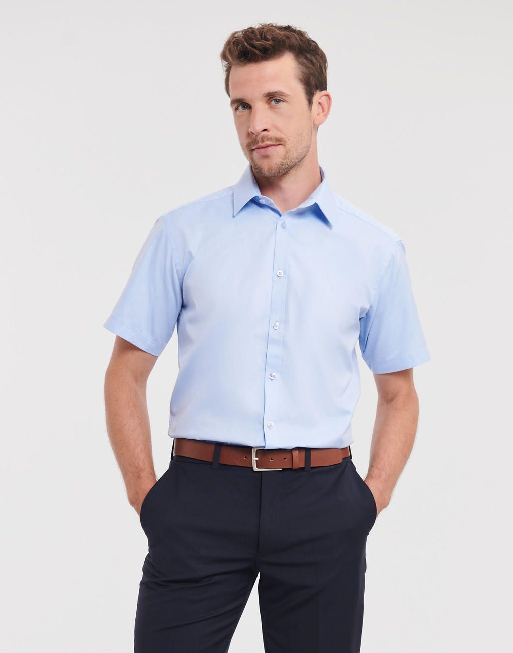 Man in a light blue short-sleeved shirt, navy pants, and brown belt. He has hands in pockets, smiling.