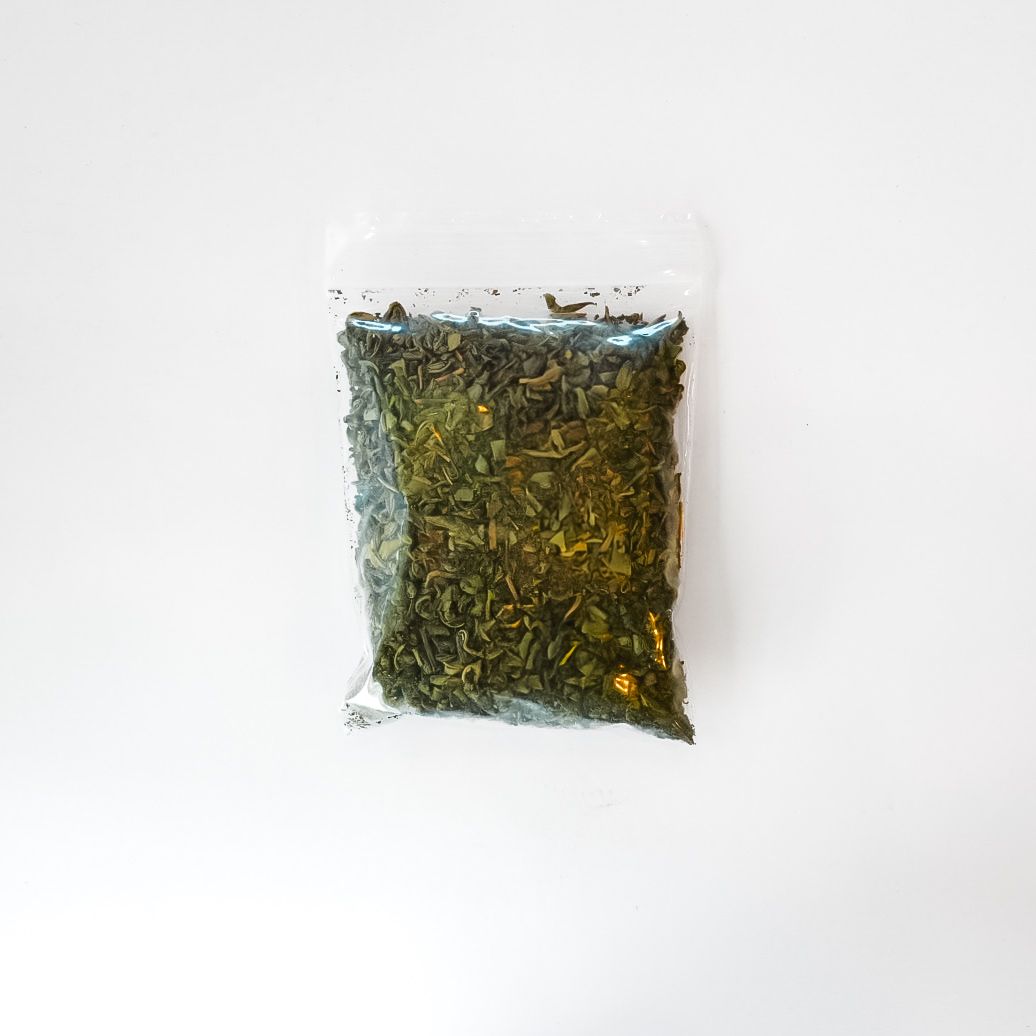 Small clear plastic bag of dried green herbs with a bright yellow label on a white background
