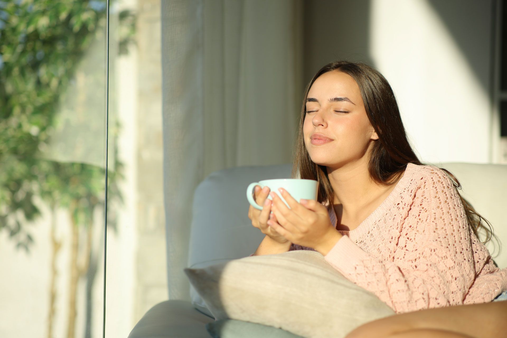 Woman relaxing on a sofa by a sunlit window, holding a mug with eyes closed.