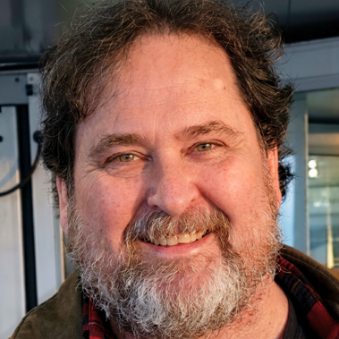 Smiling bearded man in a plaid shirt, standing indoors near a window.