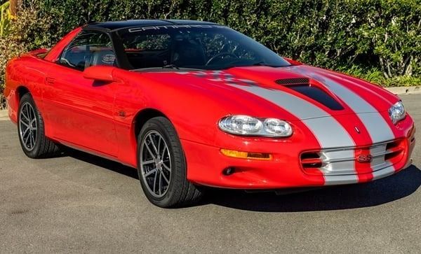 Red sports car with silver racing stripes parked on pavement under bright sunlight
