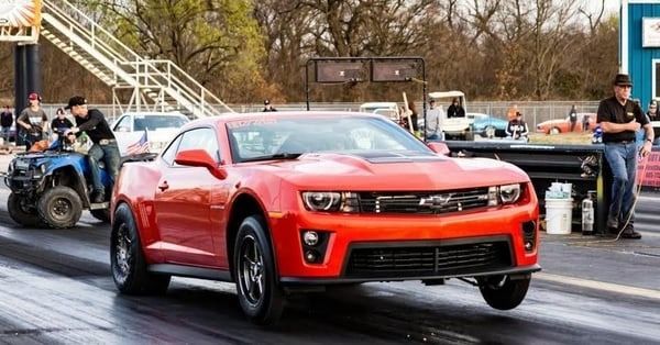 Red sports car launching on a drag strip with spectators and other vehicles in the background