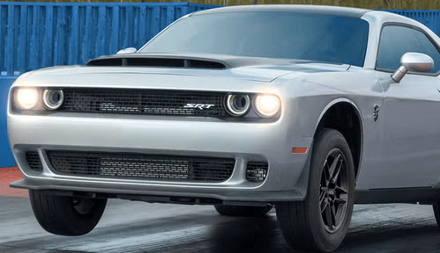 Silver Dodge Challenger driving on a wet road with headlights on, in front of a blue fence