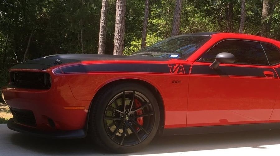 Red Dodge Challenger parked outdoors in a forest, with black racing stripes and dark tinted windows.