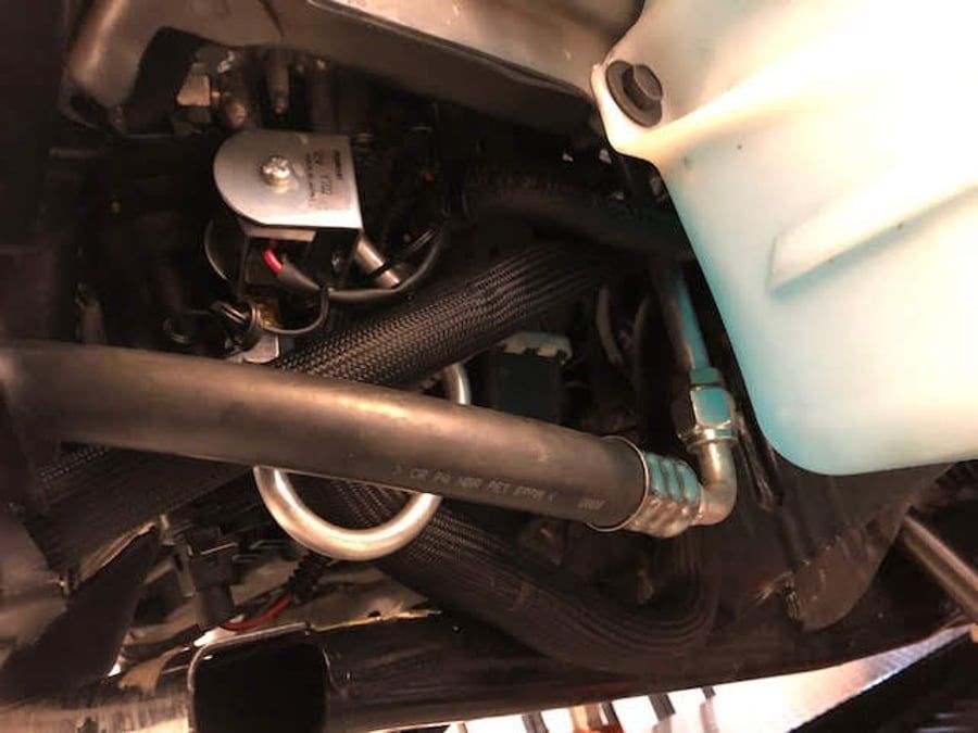 Close-up of a vehicle engine compartment with black hoses, a white fluid reservoir, and metal fittings.