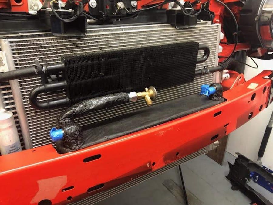 Red bumper with black oil cooler and hoses mounted on a vehicle front.