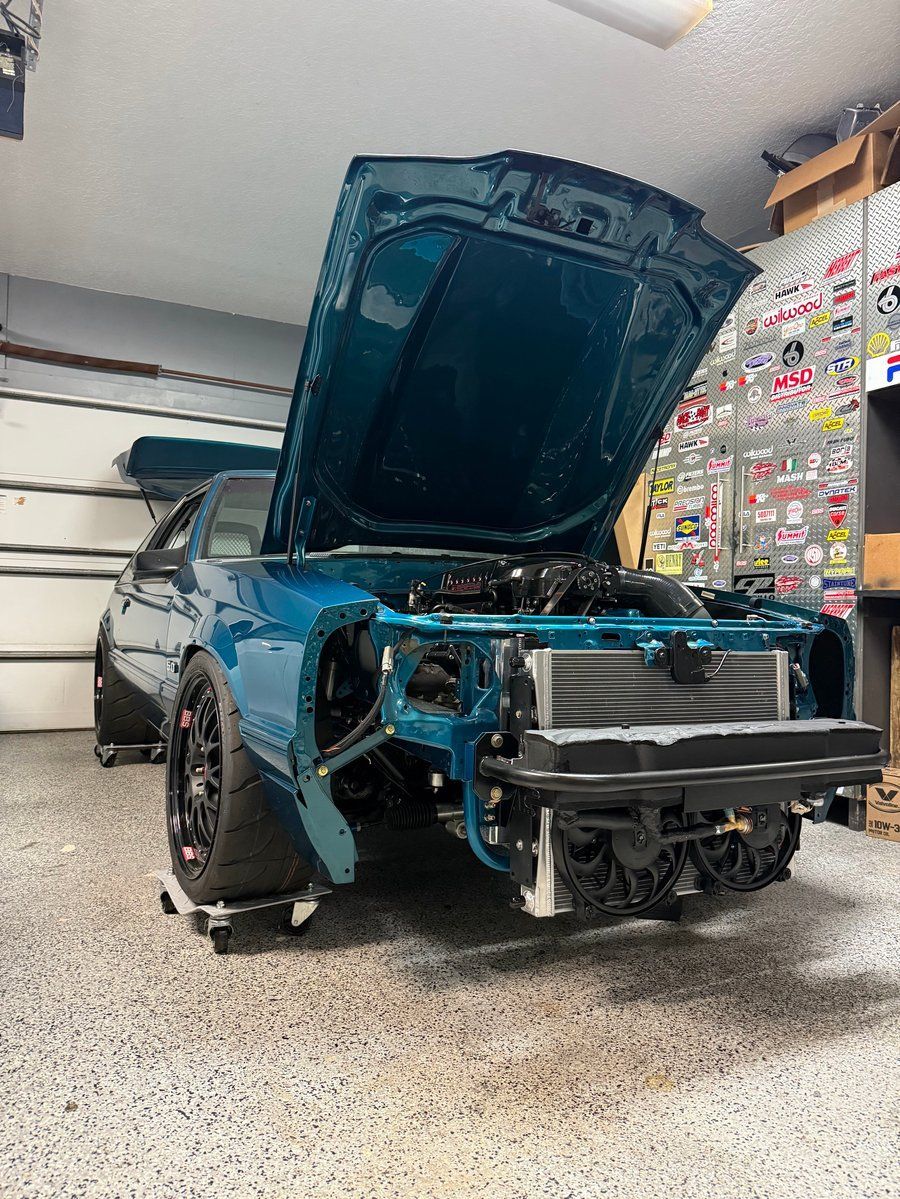 Blue car in a garage with hood open and front bumper removed, exposing the engine bay.
