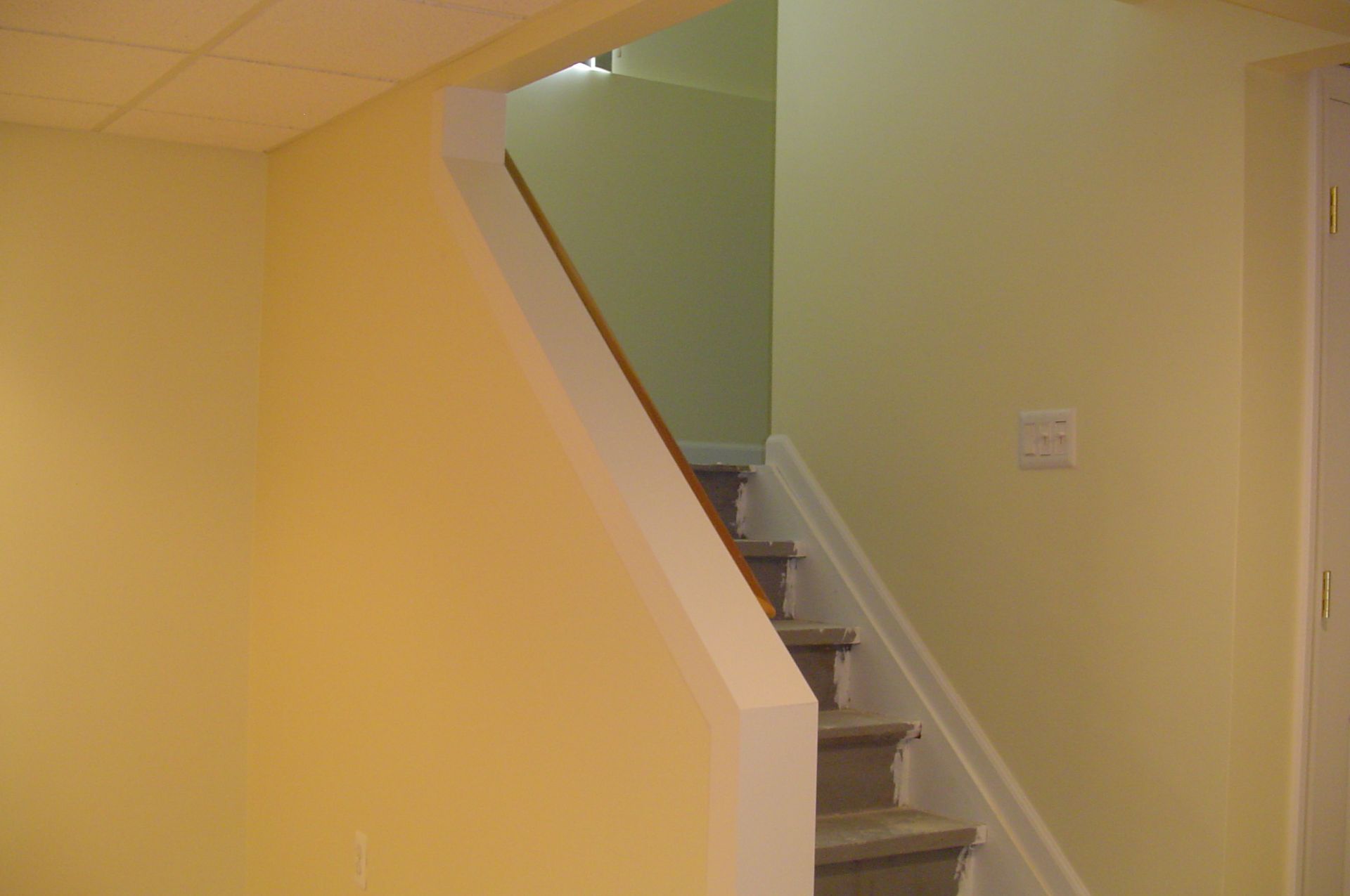 Yellow-walled interior with stairs leading up, white banister and trim, light switch, doorway.