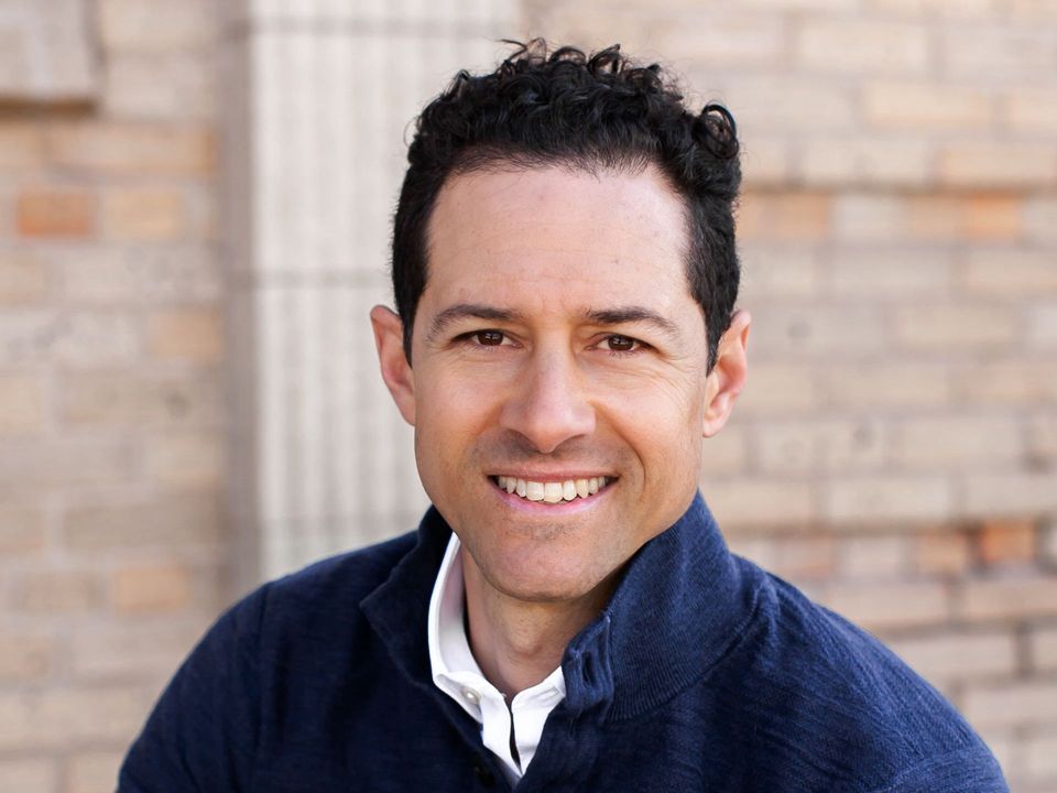 A man in a blue sweater is smiling in front of a brick wall.