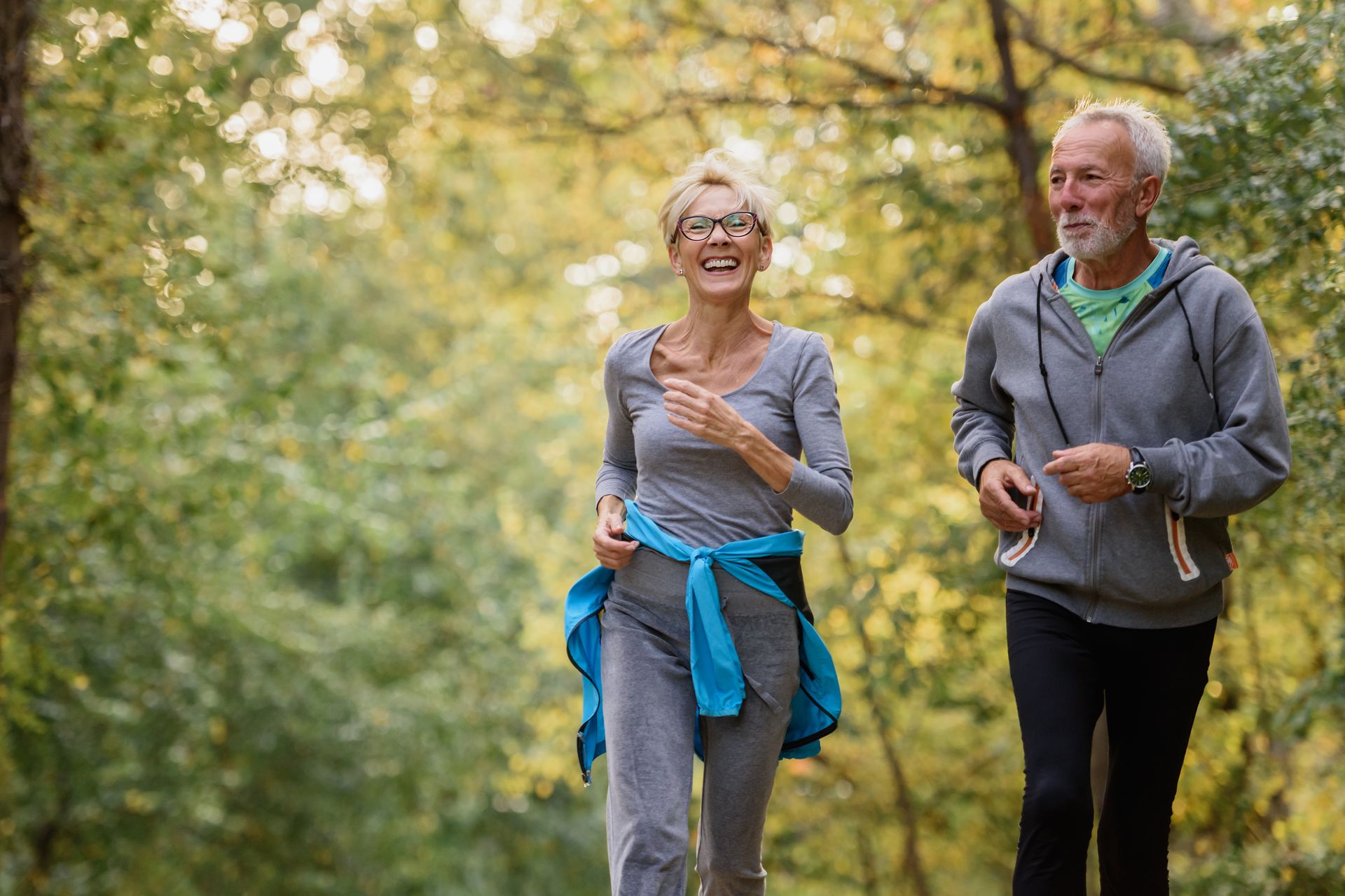 Two people running on a path through a sunlit forest, wearing comfortable athletic clothing and smiling.