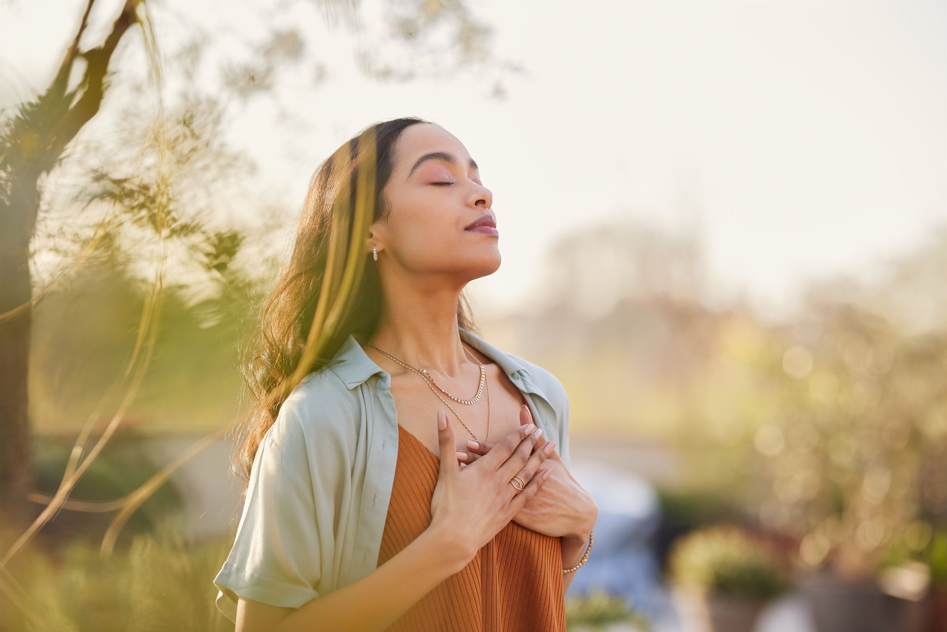A person with eyes closed, hands held over their heart, standing outside in soft, warm sunlight.