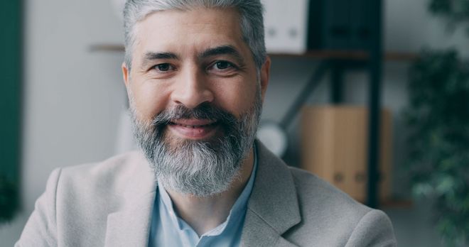 Smiling man with gray beard in a light blazer, seated indoors against a softly blurred office background