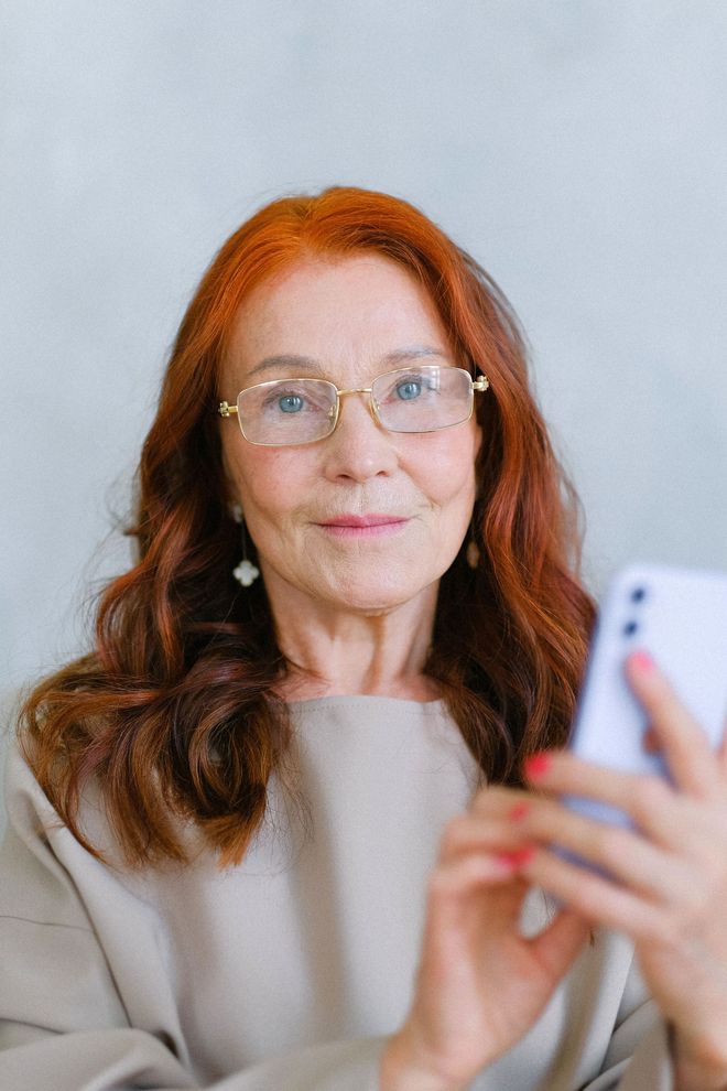Smiling woman with red hair and glasses taking a selfie on a smartphone, wearing a beige top.