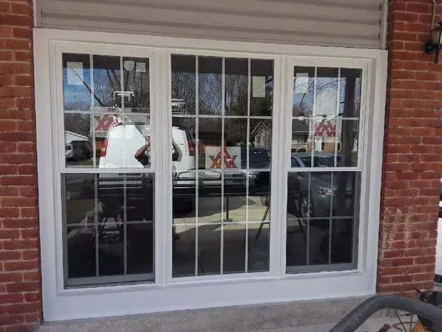GALLERY. Triple-pane window with white trim, set in a brick wall. Reflections of vehicles and trees visible.