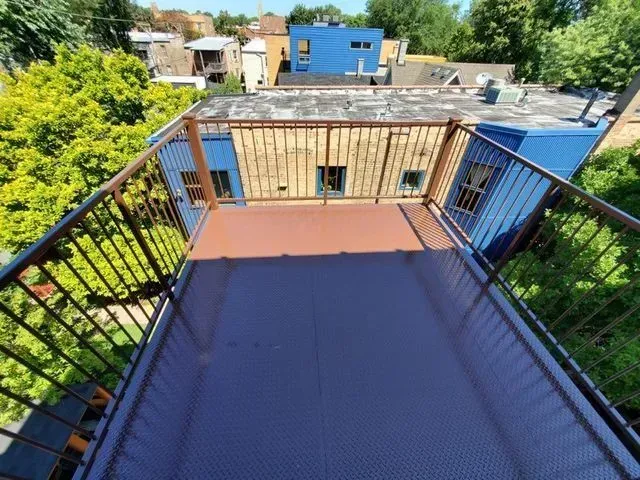 GALLERY. Overhead view of a rooftop deck with metal railings and blue accents; trees and buildings in the background.