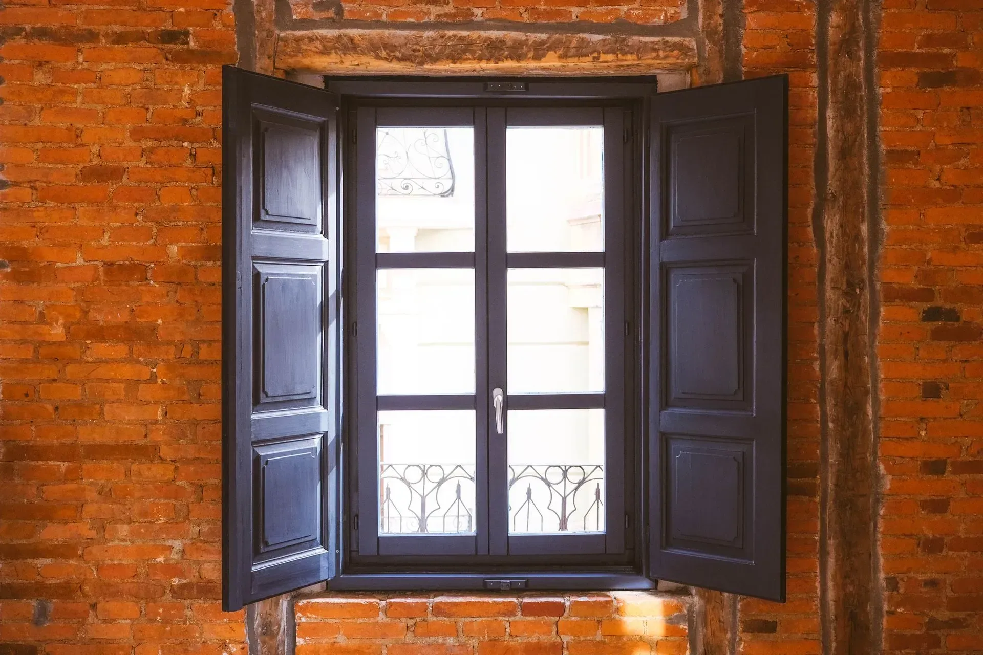 Black window with open shutters against a brick wall.