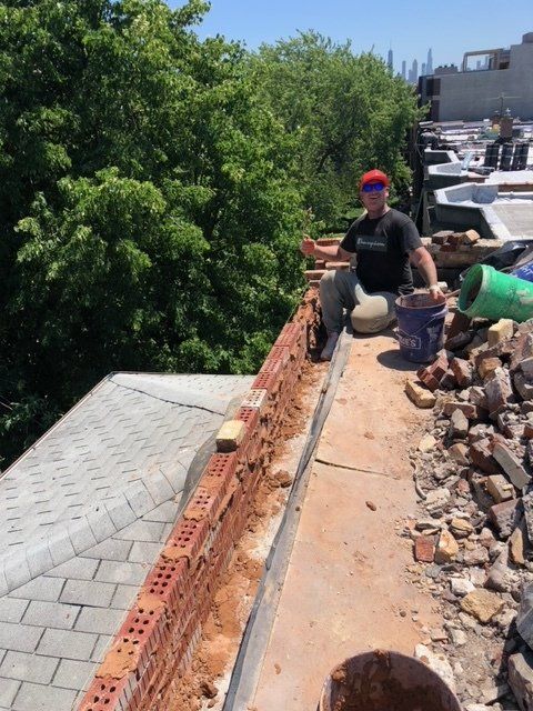 GARAGE. A person sitting on a rooftop building a brick wall with supplies nearby on a sunny day.