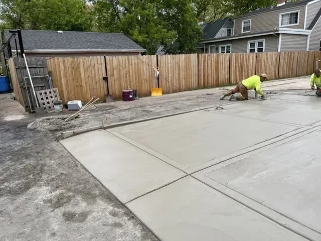Gallery. Workers in safety vests finishing a newly poured concrete driveway next to a wooden fence.