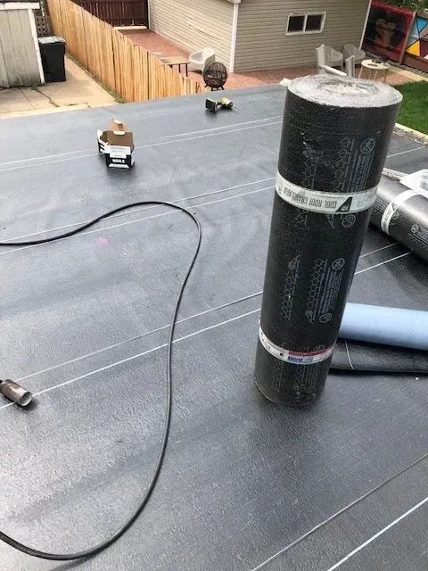 Gallery. Roll of roofing material on a flat roof with lines, tools, and a wooden fence in the background.