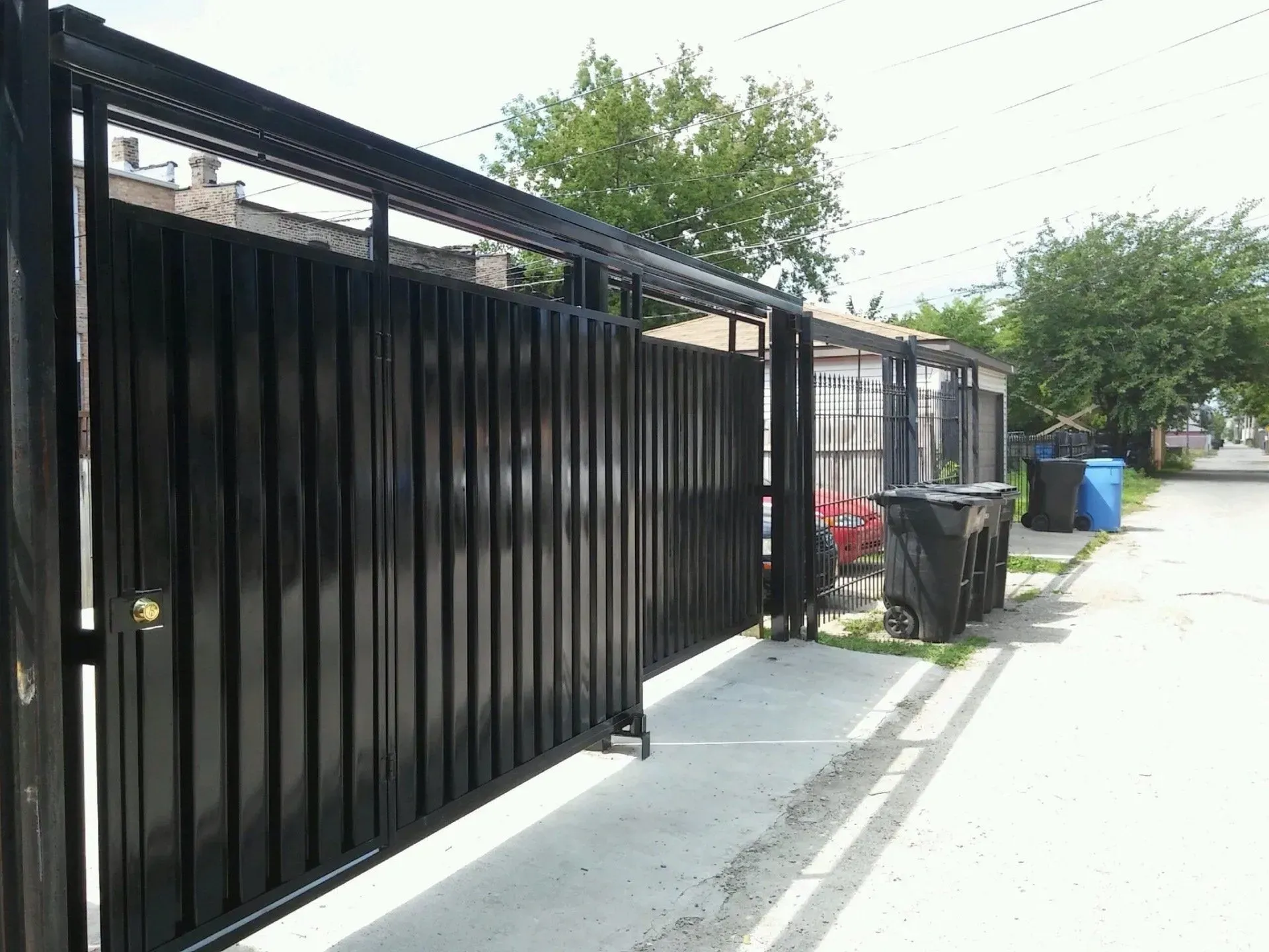 Gallery. Black metal gate and fence along a sidewalk, with trash bins visible.
