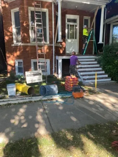 Workers renovating a red brick house with porch and steps. Equipment and materials are scattered on the sidewalk.