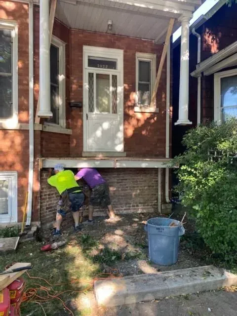 Two workers repair a brick porch.  One wears a neon vest.  The porch has support beams and a white door.