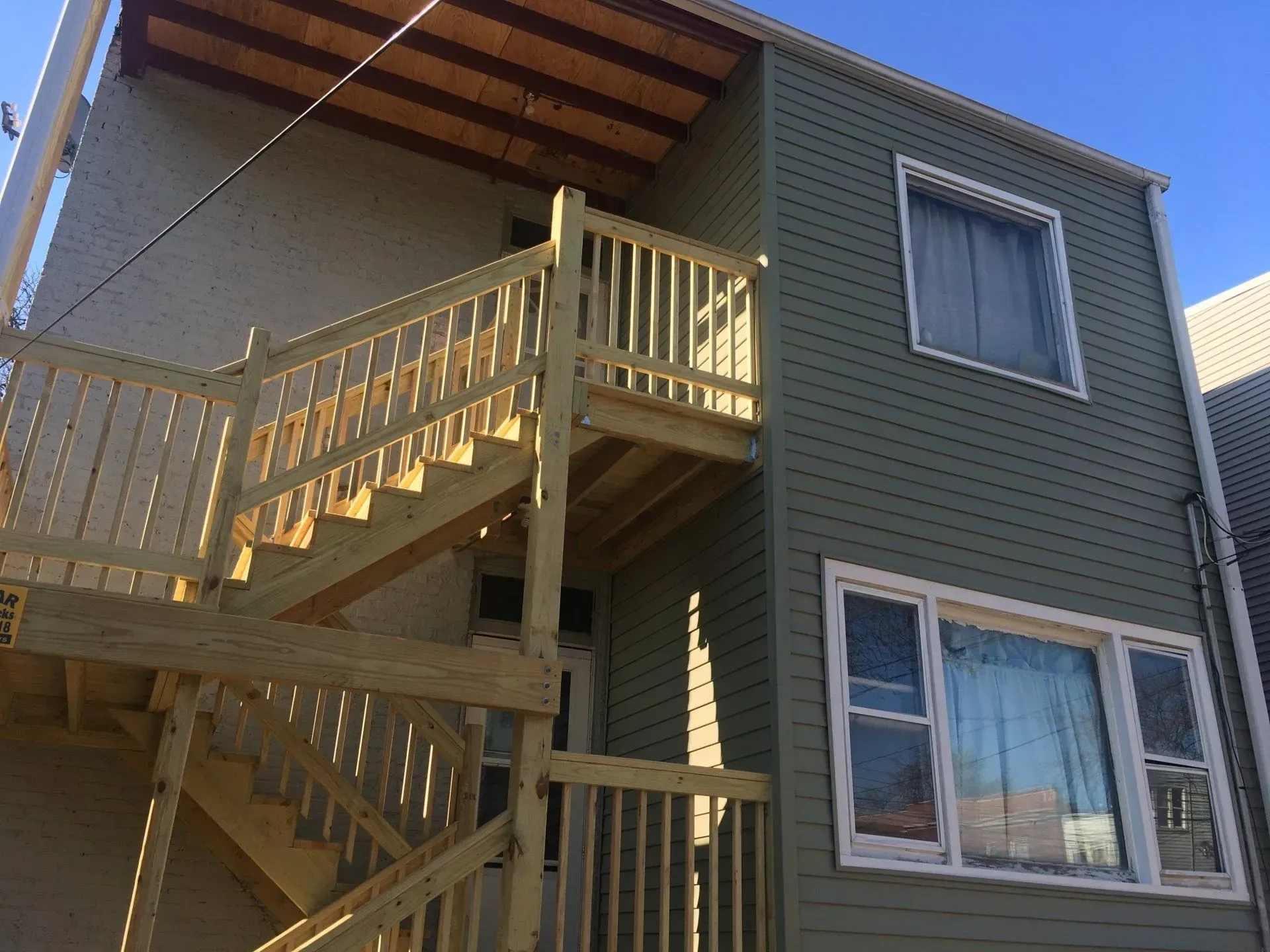 Gallery. Exterior of a two-story building with wooden stairs leading to a deck. Green siding and a blue sky are visible.