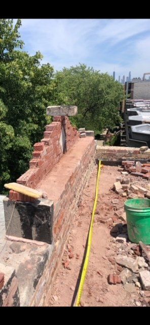 GARAGE. Brick chimney under construction on a rooftop, green bucket, trees and a blue sky in the background.