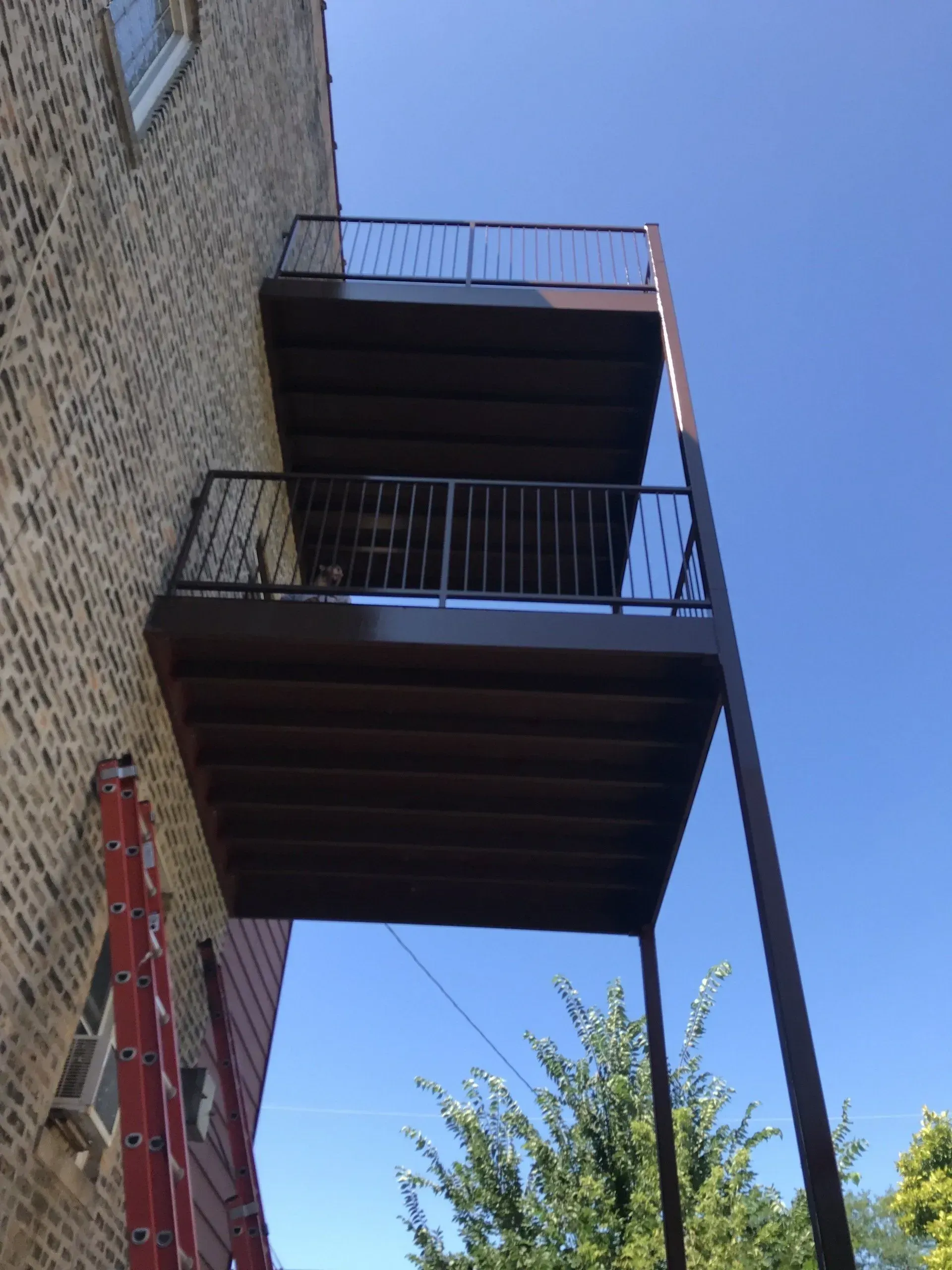 Gallery. Two-tiered brown deck balconies with black railings attached to a brick building, supported by a brown pole.